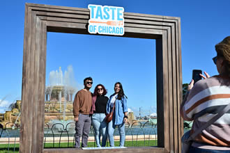Taste of Chicago Grant Park (Person taking a photo of three people standing under a frame with the Taste of Chicago logo at the top and Buckingham Fountain in the background)